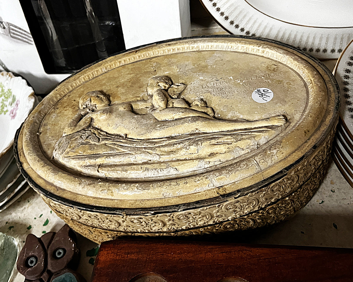 Oval decorative piece with figurative relief, an inscription and ornate sides, photographed among auction objects.